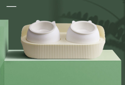 Two Ceramic Food and Water Bowl Stations for Cats sitting on top of a green plant. (Brand: CJdrop)